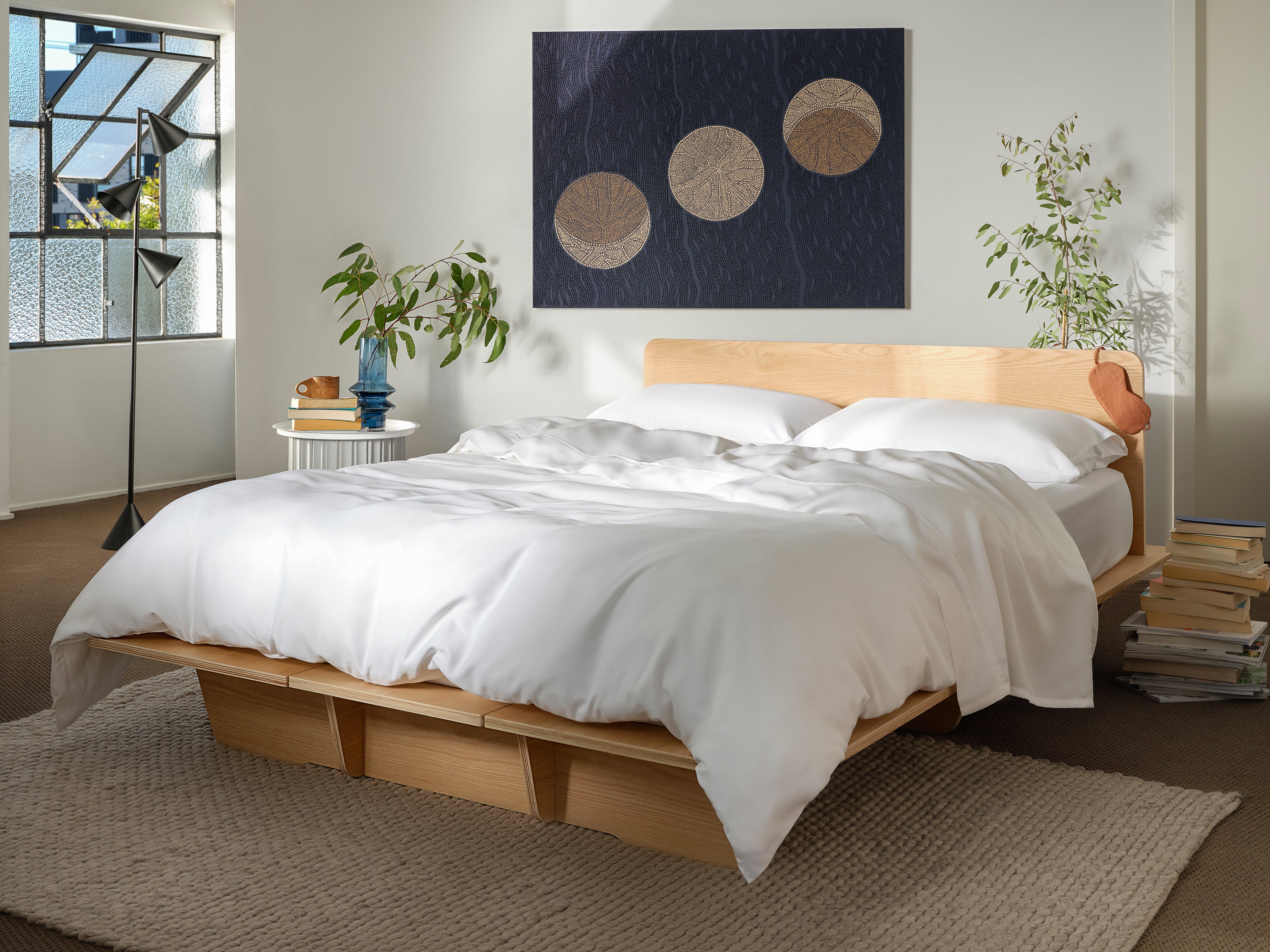 Minimalist bedroom featuring a light wood platform bed with white bedding, large abstract artwork, and plants on a neutral rug.