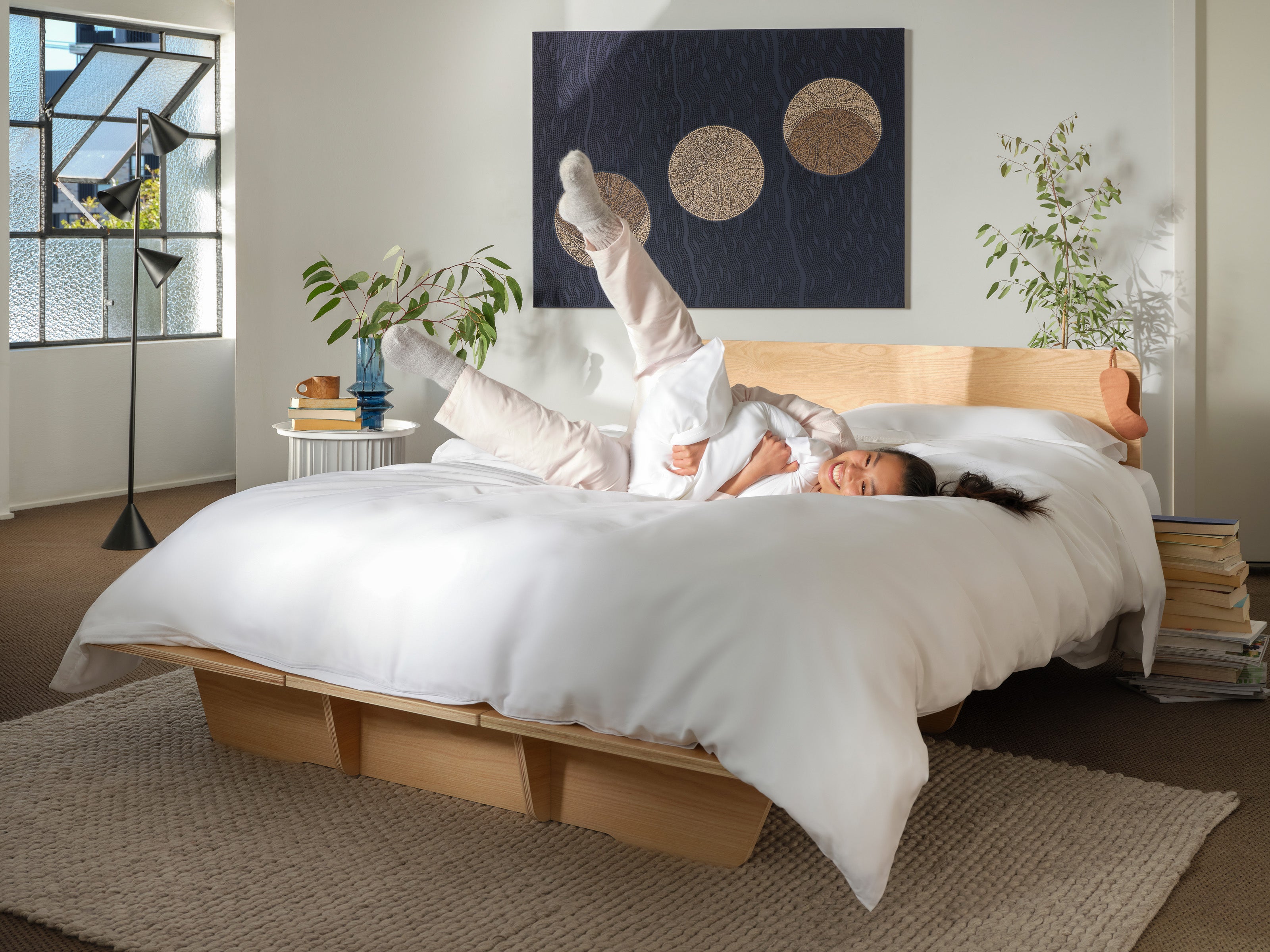 A woman playfully relaxes on a light wood bed with white bedding. The room features indoor plants, a rug, and abstract wall art.