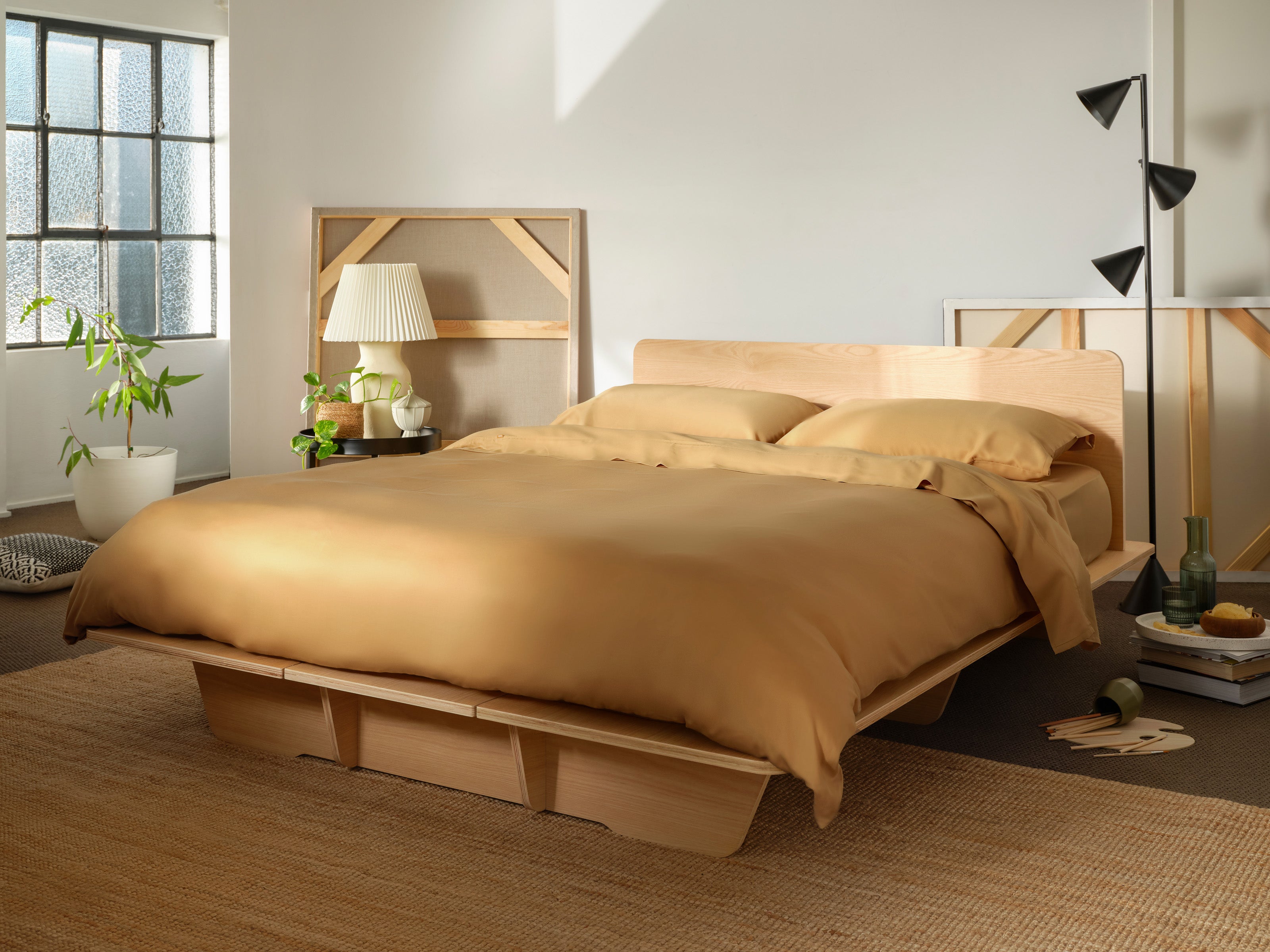 Minimalist bedroom with a light wood bed frame and mustard bedding. Indoor plant, lamp, and window create a serene atmosphere.