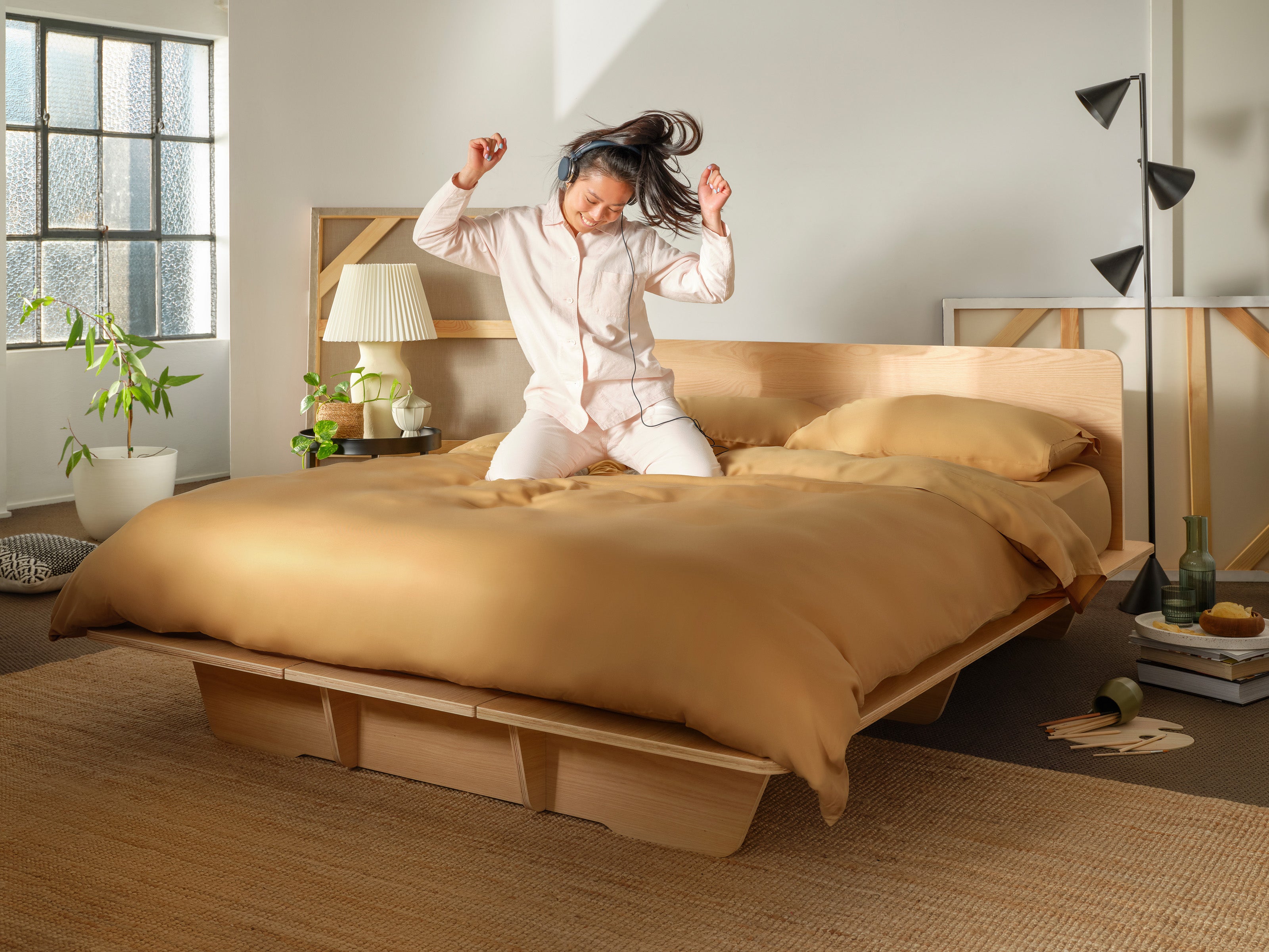 A woman in pyjamas joyfully jumps on a bed with a wooden frame and mustard bedding. A lamp and plant adorn the nightstand; natural light streams in.