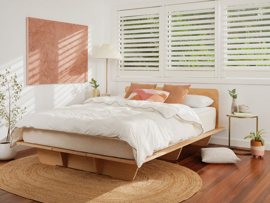 Minimalist bedroom with a wooden platform bed, white bedding, three cushions, a round rug, and a potted plant. Sunlight filters through shutters.