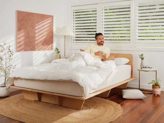 Man relaxing in a modern bedroom with a wooden bed, white bedding, and natural decor. Large windows with shutters; plants and art adorn the space.