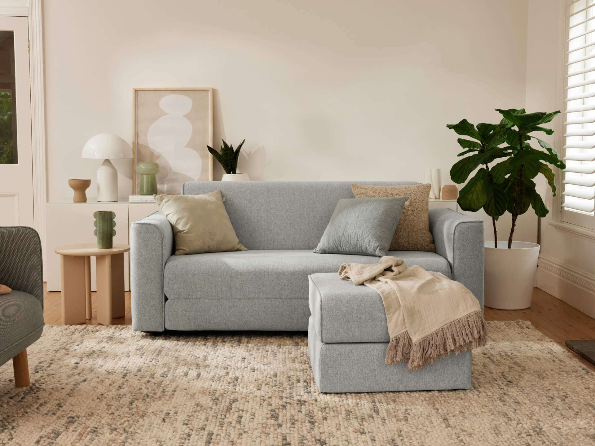 Light grey sofa with cushioned ottoman in a cosy living room. Neutral decor, indoor plant, art, and a textured rug enhance the setting.