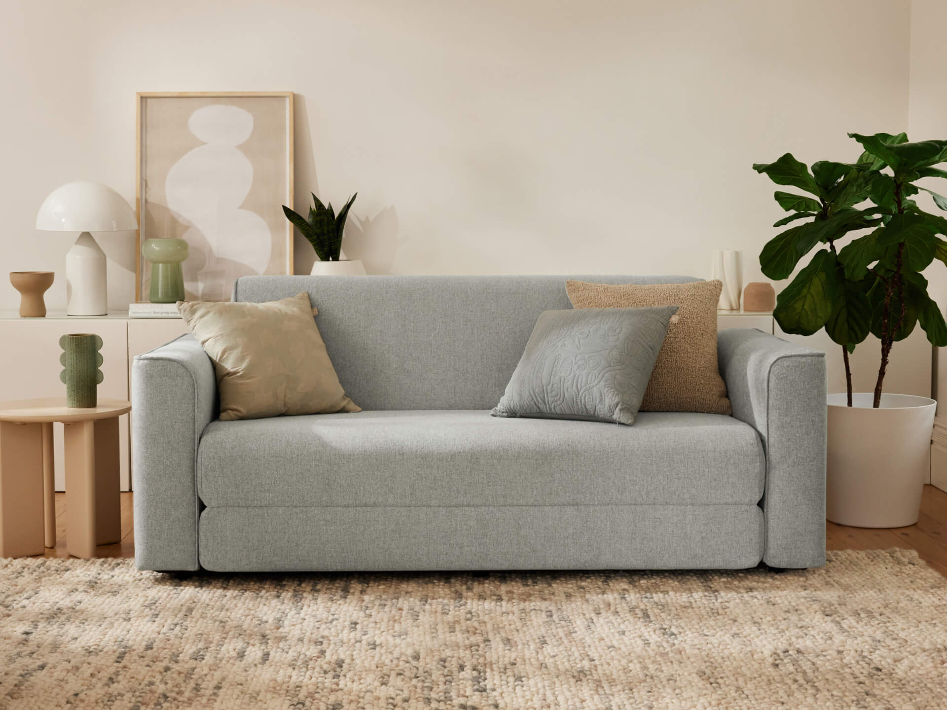 Light grey sofa with multiple cushions in a cosy living room. Neutral decor, indoor plant, side table, and modern art in the background.