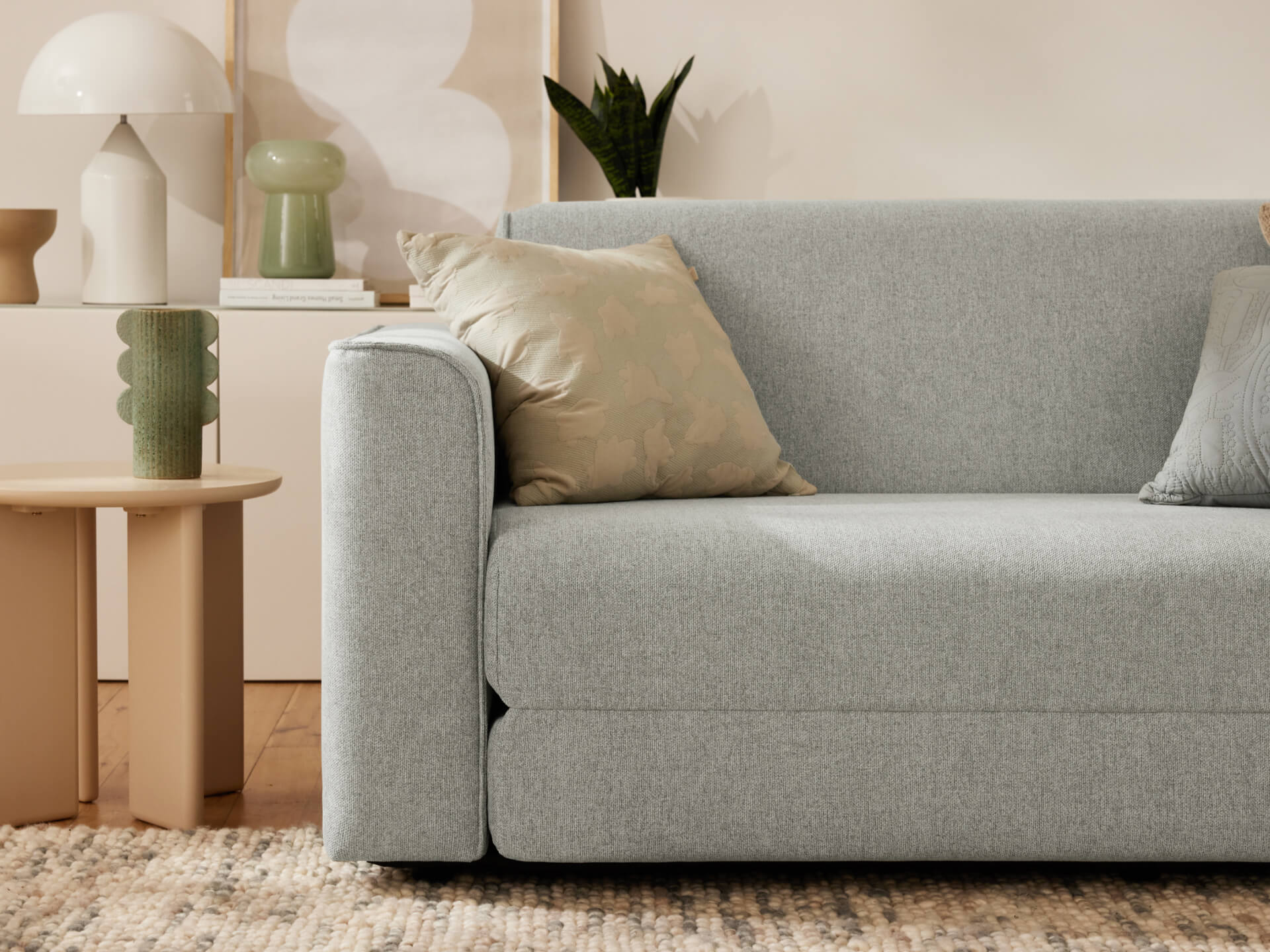 Light grey sofa with two cushions in a cosy living room. Beige side table with decor and plants in the background. Warm, neutral tones.
