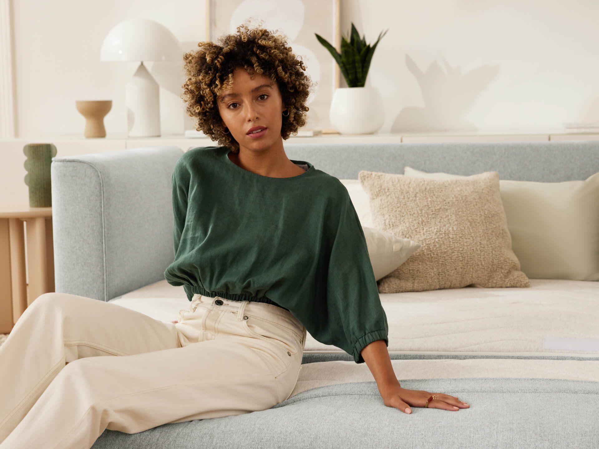Woman seated on light grey sofa with textured cushions and neutral decor in the background. She wears a dark green top and light trousers.