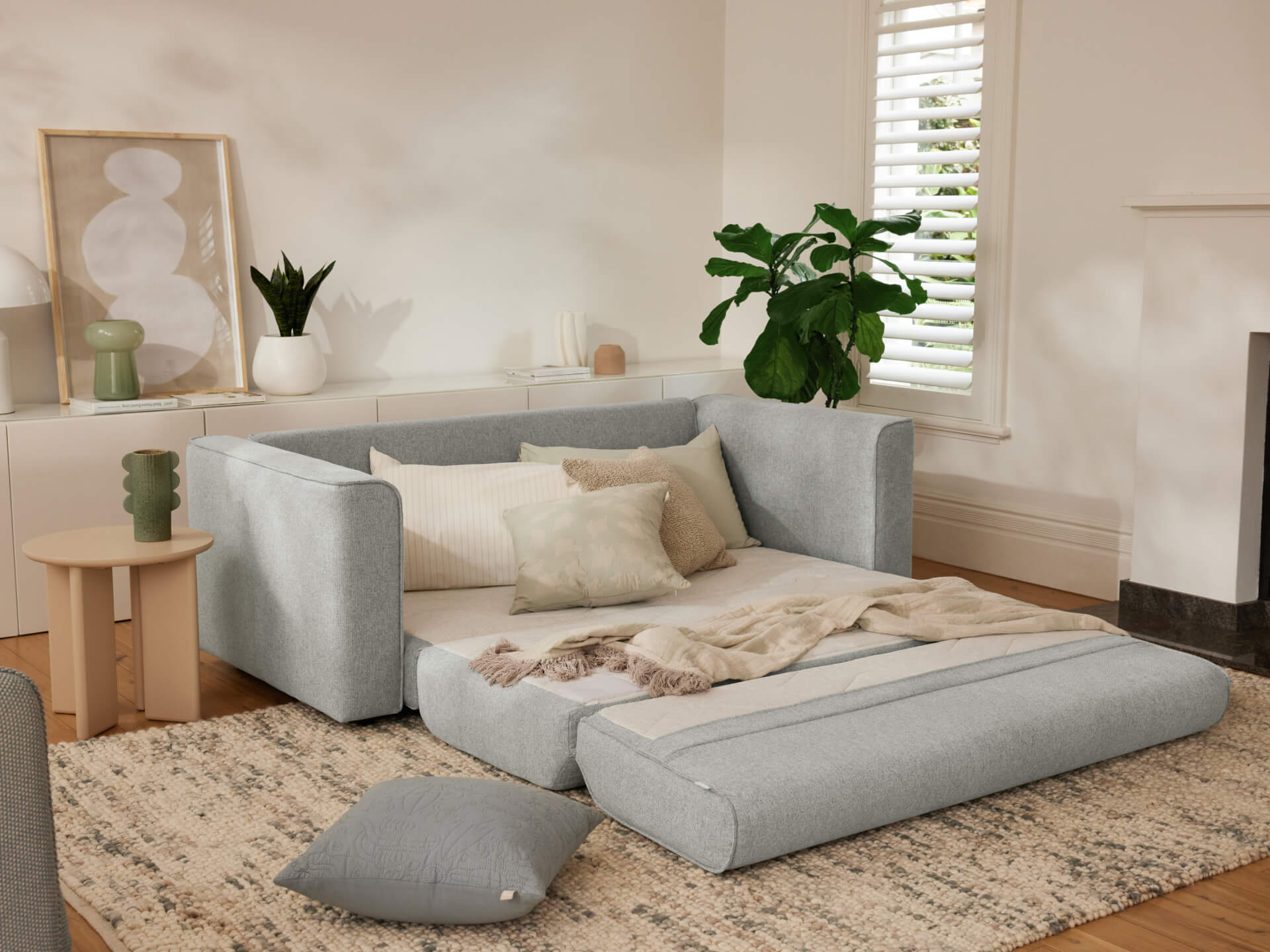 Light grey sofa bed extended in living room with beige cushions, wooden side table, green plant on cabinet, and abstract art on wall.