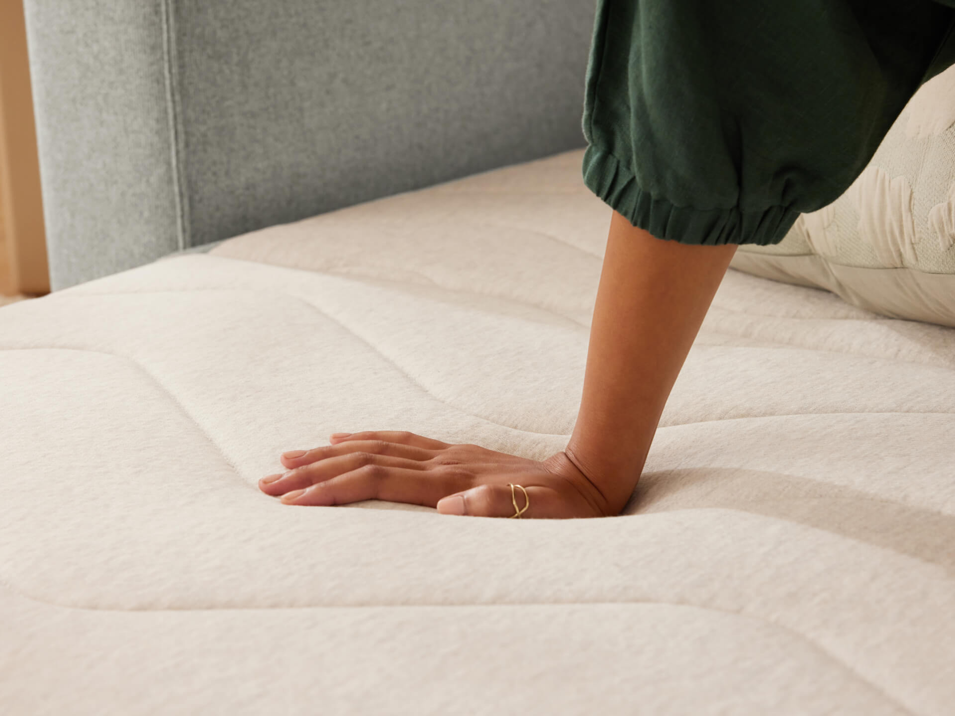 Person pressing hand onto a beige mattress with a quilted texture, next to a green cushioned headboard.