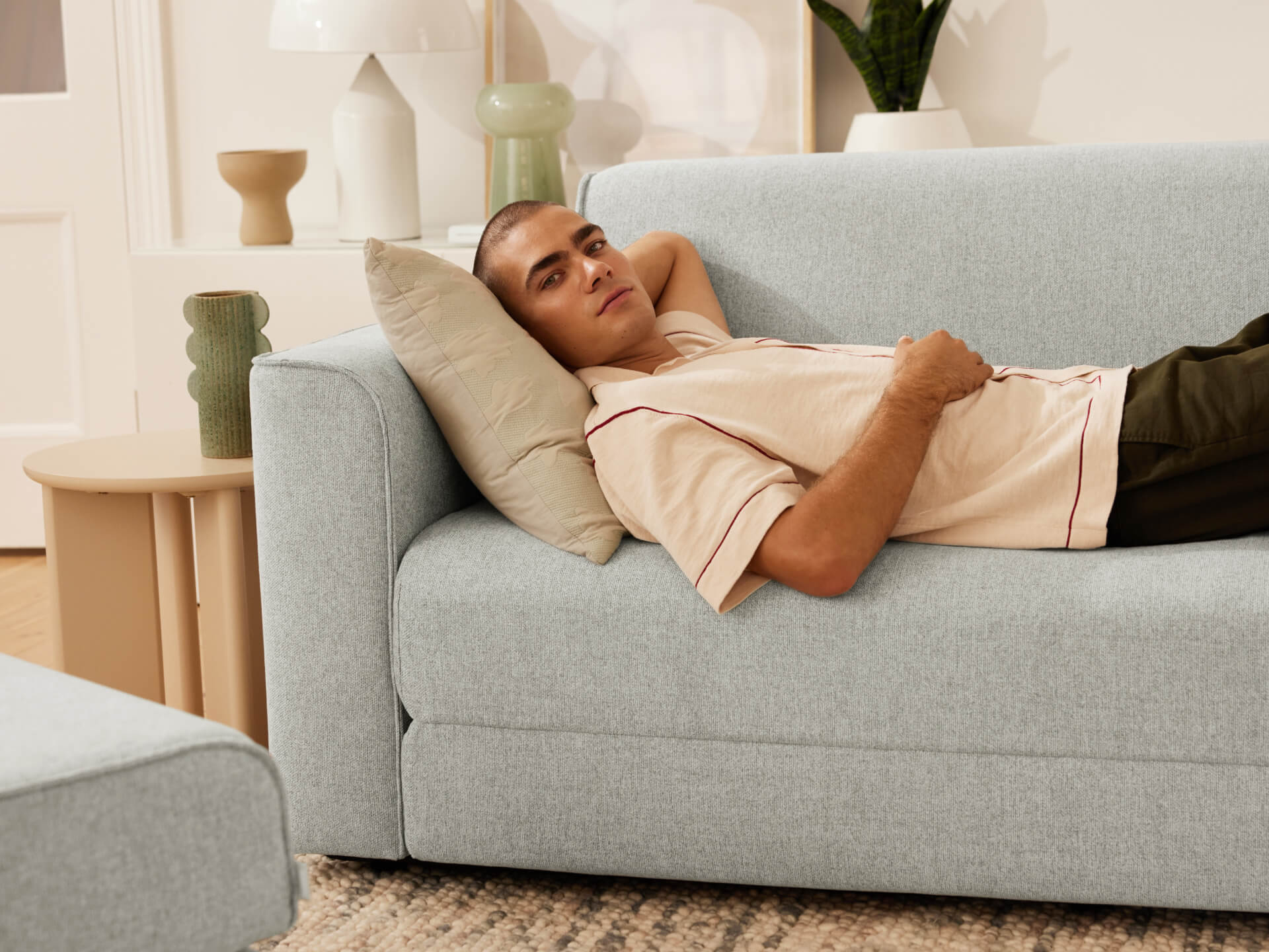 Man in beige shirt lounging on a light grey sofa with a cushion, set in a modern living room with decorative vases and plants.
