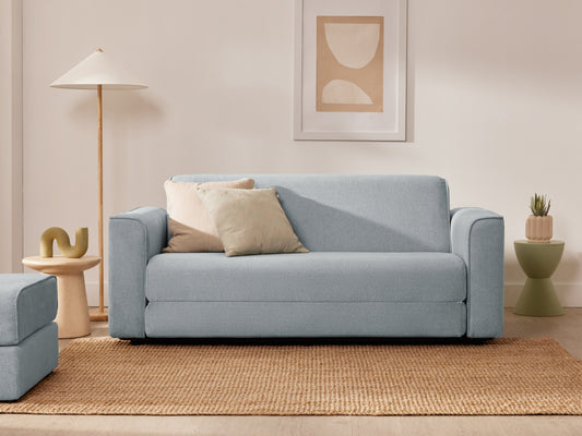 Light blue sofa with beige cushions on a woven rug. Nearby are a tall floor lamp, green side table, plant, and abstract wall art.