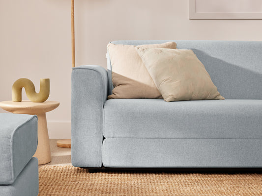 Light blue sofa with beige cushions on a woven rug. A wooden side table holds a ceramic decor piece. Soft, neutral room tones.