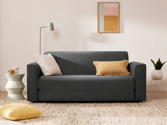 Dark grey sofa with beige and yellow cushions, beside a light grey side table. A modern floor lamp stands nearby, with art on the wall and a rug underneath.