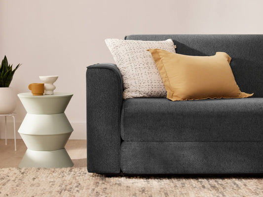 Dark grey sofa with beige and white cushions, next to a unique tiered side table holding mugs. A textured rug and plant are in the background.