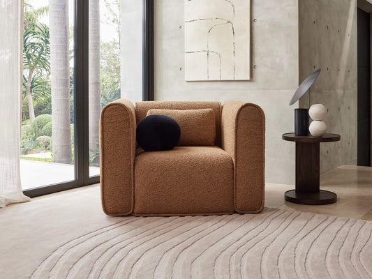 Beige armchair with rounded edges, black and beige cushions. Dark wood side table with decorative objects. Abstract art, large window, light rug.