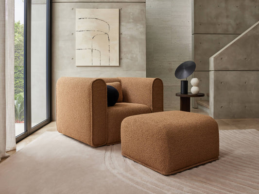 Modern living room with a brown upholstered armchair and matching ottoman. Concrete walls feature abstract art; large windows allow natural light.