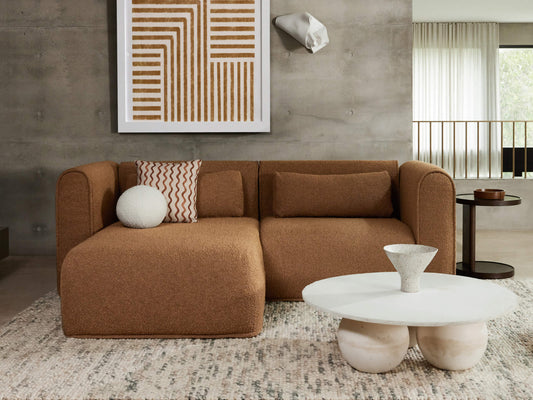 Stylish living room with a brown sofa, textured cushions, round white coffee table, abstract wall art, and earthy-toned decor.