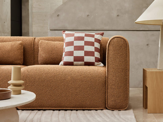 Brown textured sofa with checkered cushion, beside a beige lamp on a wooden side table. Rug and ceramic decor in foreground. Modern interior design.