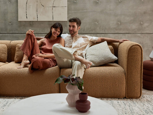 A couple sits on a beige, textured sofa with cushions, beside a white round coffee table with vases; a concrete wall and abstract art behind.