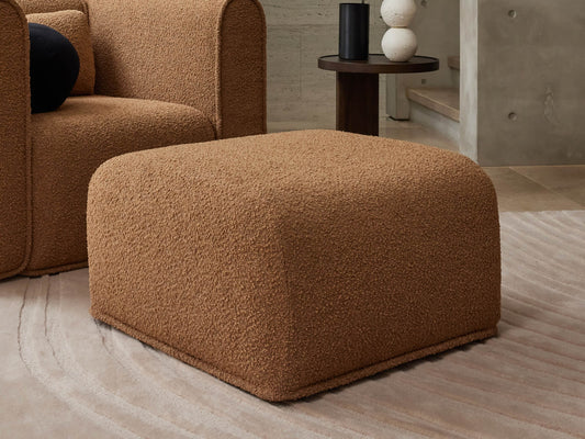 Brown textured ottoman on a light beige patterned rug, with a matching chair and dark round side table in the background.
