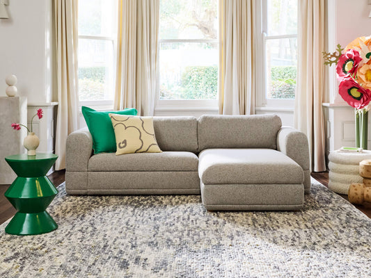 A light grey sectional sofa with green and beige cushions sits on a patterned rug. Nearby are a green side table and large floral decor.