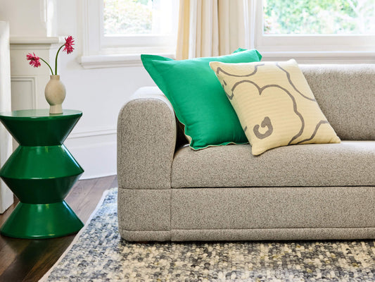 Light grey sofa with green and patterned beige cushions, next to a green side table with a vase and pink flowers, on a multicolour rug.