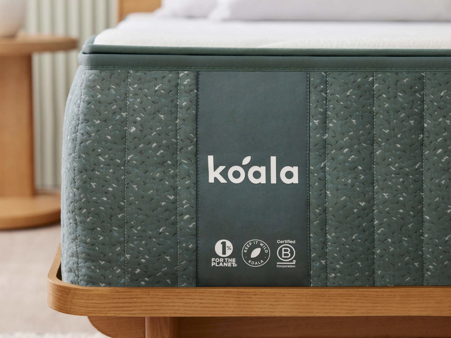 Teal mattress with "koala" branding on a wooden bed frame, featuring eco-friendly and certified icons. Cream pillow and sheets partially visible.