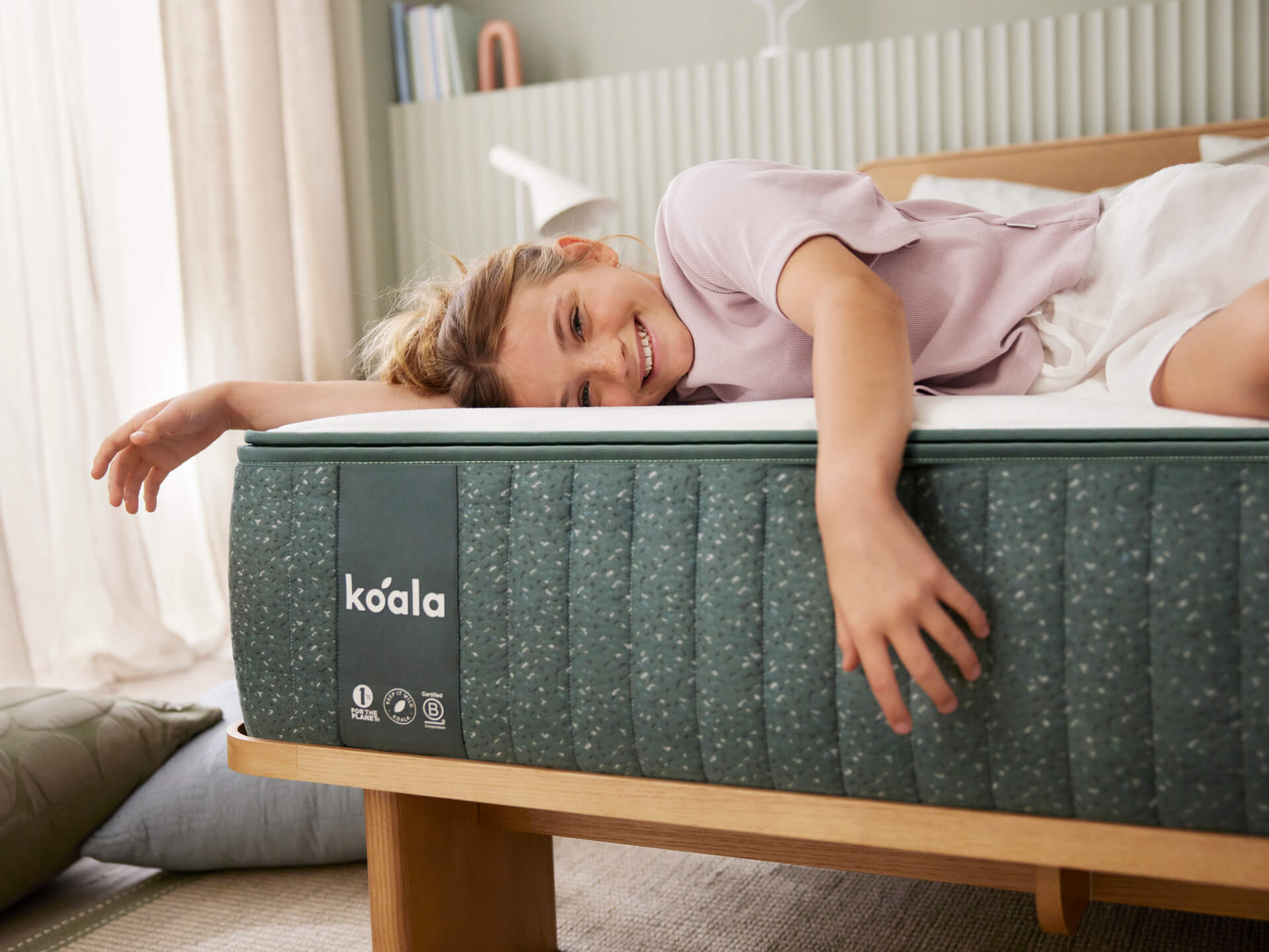 Girl smiling and lounging on a Koala mattress with a grey quilted cover and wooden bed frame, in a light-filled, cosy bedroom.