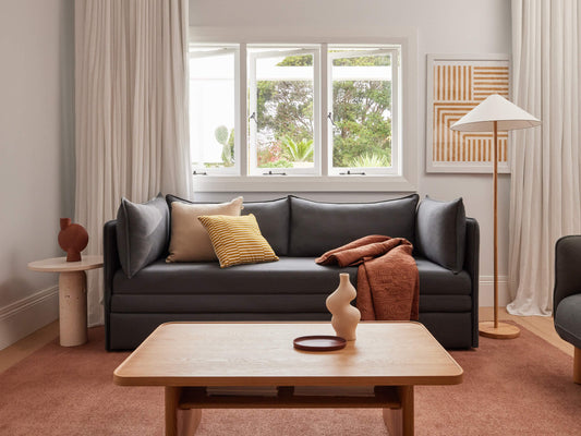 Minimalist living room with a dark grey couch, mustard and rust cushions, light wood coffee table, tall lamp, and abstract art. Neutral tones.