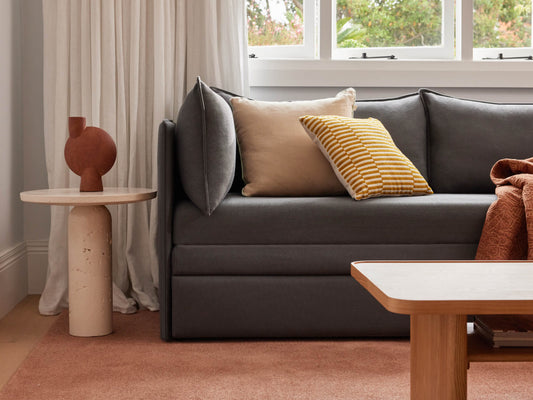 Dark grey sofa with beige and yellow-striped cushions, beside a round side table with a brown vase. Light wood coffee table and peach rug.