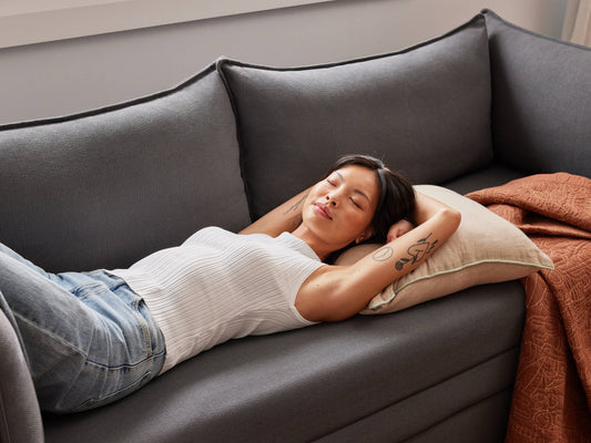 Woman relaxing on a dark grey couch with a light cushion and rust-coloured throw, wearing a white top and jeans, arms behind head.