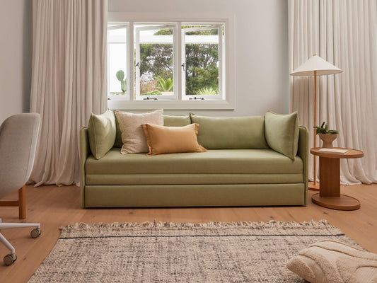 Green sofa with cream and orange cushions in a living room. Cream curtains, light wood lamp and side table, grey office chair, and patterned rug.