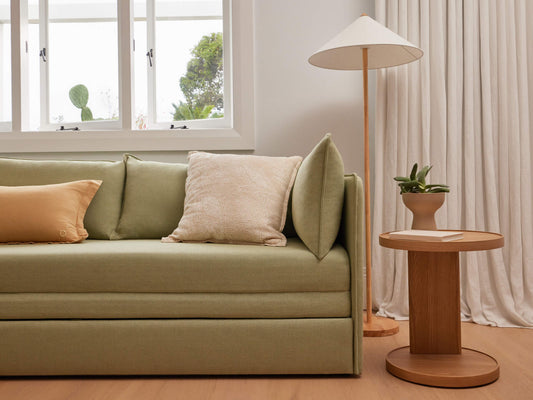 Minimalist living room with a light green sofa, beige and yellow cushions, wooden side table, floor lamp, and potted plant by a window.