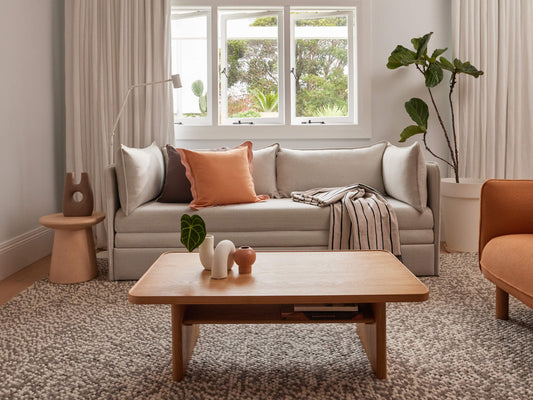 Modern living room with a light grey sofa, peach pillow, striped throw, wooden coffee table, grey rug, indoor plant, and white curtains.