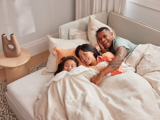 Family lying on a cream corner sofa, covered with a beige blanket. A small round wooden table with a decorative vase is beside them.