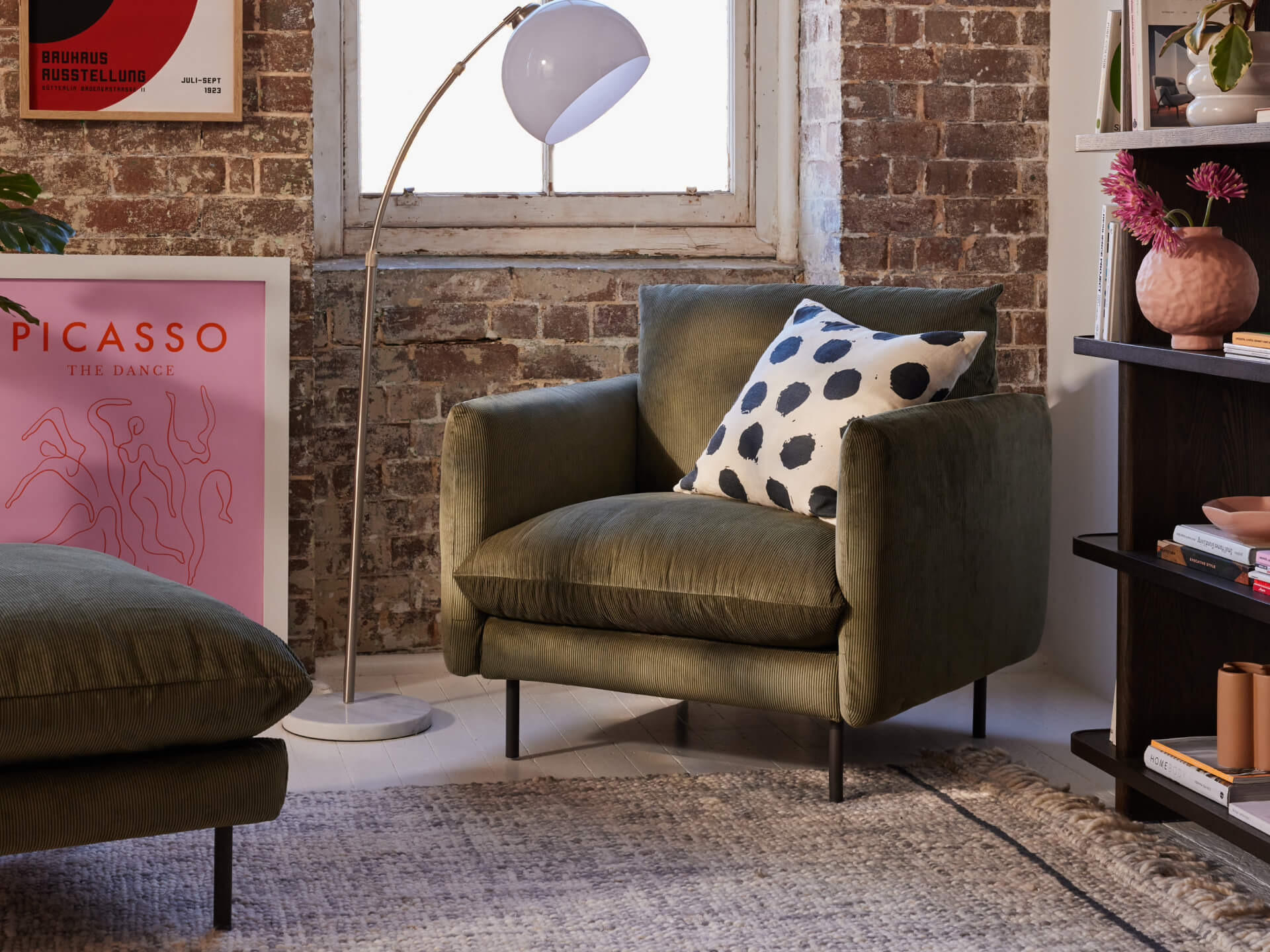 Olive green armchair with black polka dot cushion, against an exposed brick wall, next to a white floor lamp and decorative items.