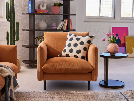 Orange armchair with black polka dot cushion next to a round black side table with pink flowers. Shelving and décor in background. Light-filled room.