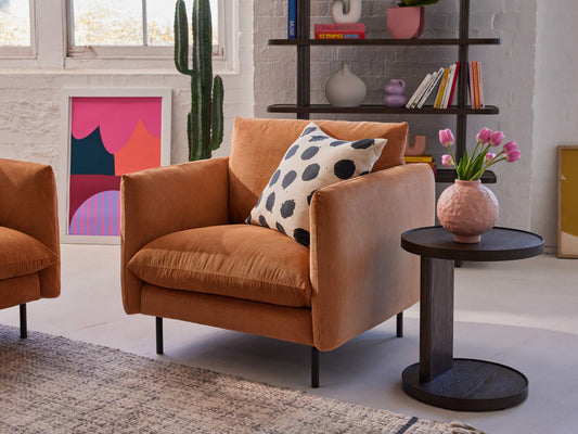 Cosy brown armchair with a black-and-white spotted cushion beside a round dark wood side table with pink flowers in a vase.