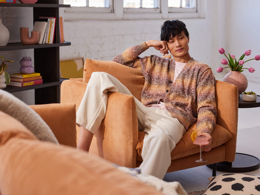 Person in a multicoloured cardigan and beige trousers reclines on a light brown armchair, holding a wine glass. Nearby, a vase holds pink flowers.