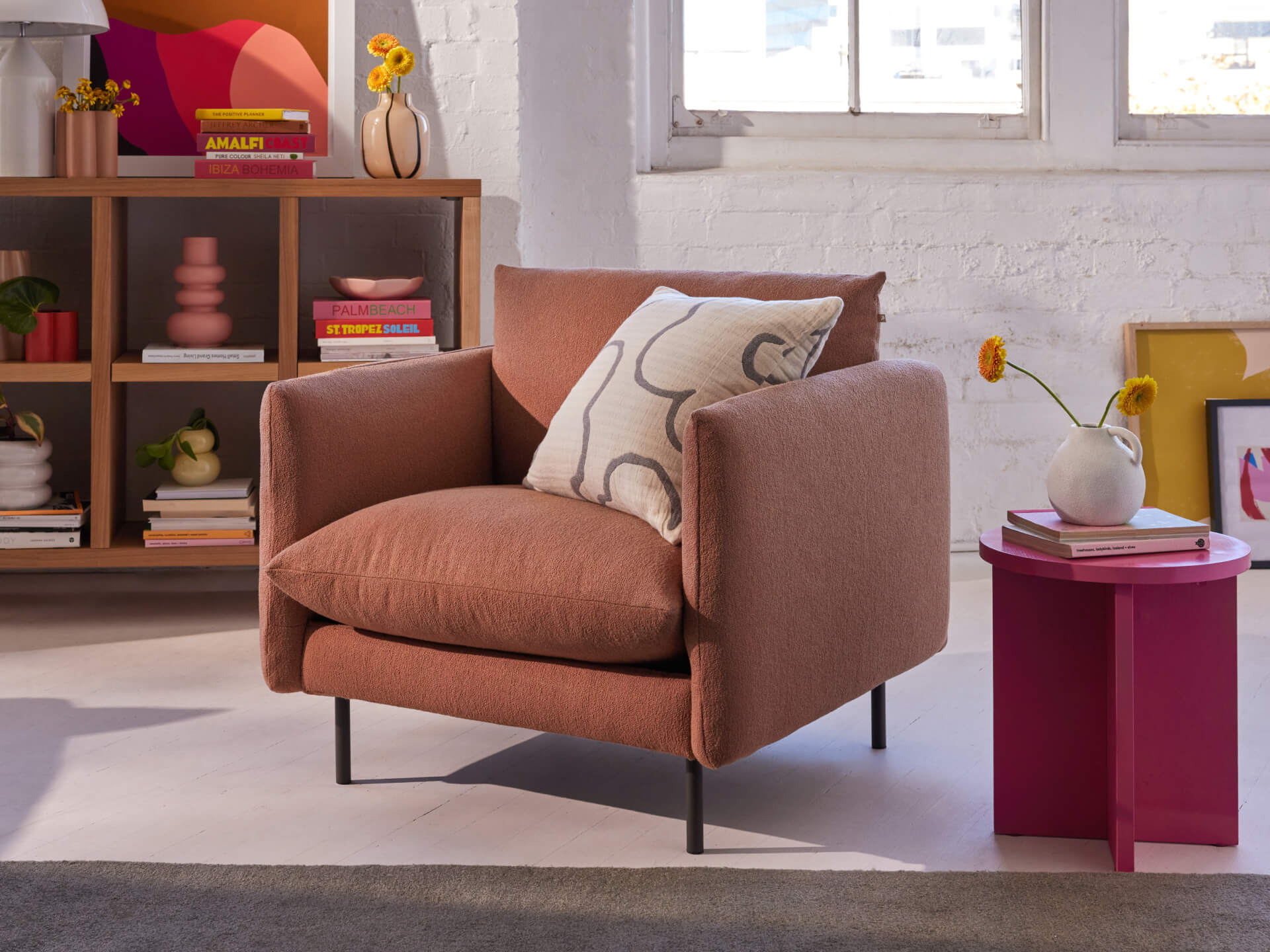 Rust-coloured armchair with a patterned cushion, beside a pink round table with a vase, against a white brick wall and shelf background.