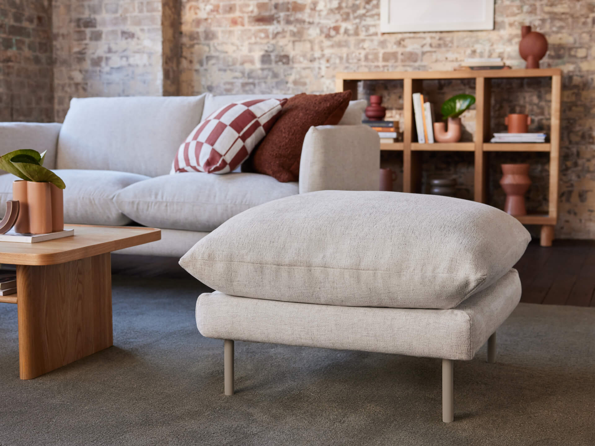 Modern living room with a grey sofa and ottoman, wooden coffee table, decorative cushions, and exposed brick wall with a bookshelf.