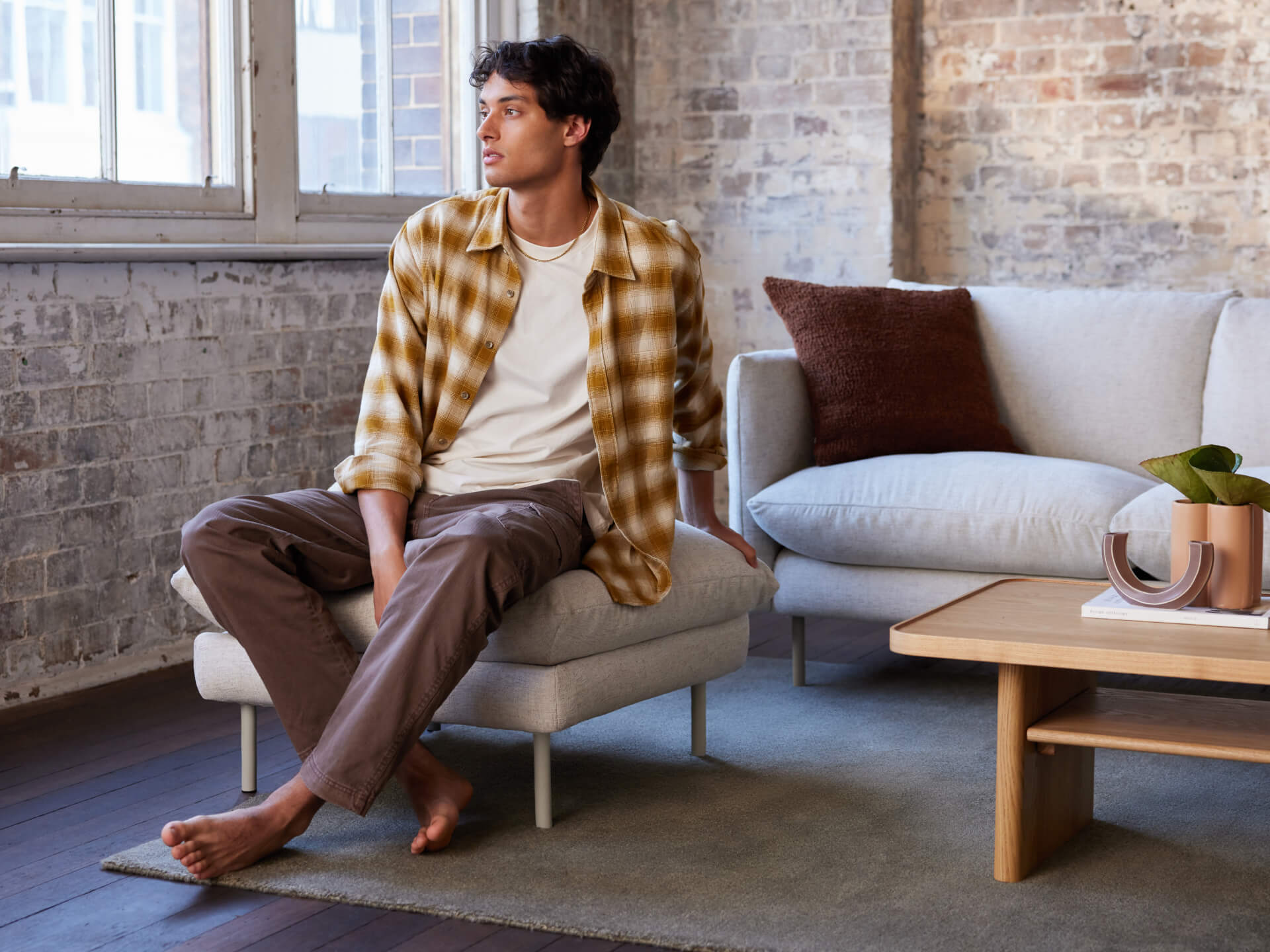 A person sits on a light grey ottoman in a stylish loft with exposed brick walls, near a soft grey couch and a wooden coffee table on a rug.