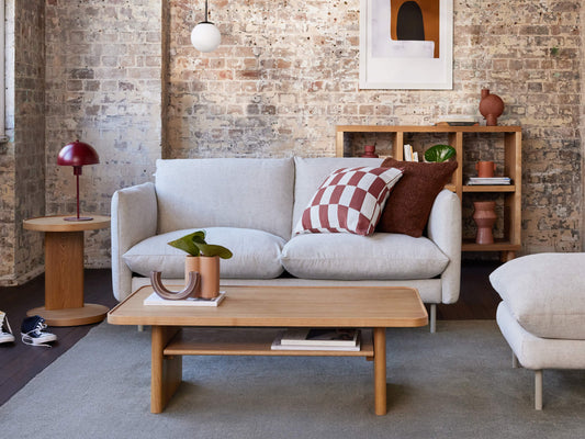 Cosy living room with light grey sofa, wooden coffee table, brick wall, and modern decor. Red and white cushions add a stylish touch.