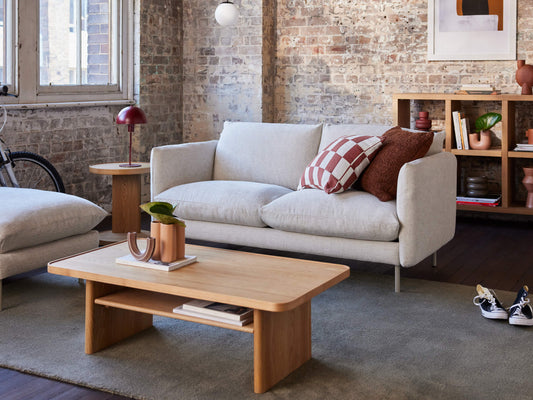 Modern living room with a light grey couch, wooden coffee table, bookshelves, decorative cushions, and large windows. Exposed brick walls add texture.