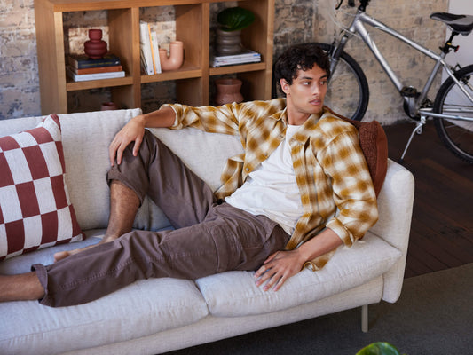 Man lounging on a light grey couch, wearing a yellow check shirt and brown trousers. Brick wall, shelf with decor, and a bicycle in the background.