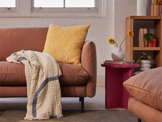 Living room with a peach sofa featuring a yellow cushion and a cream throw. Nearby, a pink side table holds a white vase with yellow flowers.