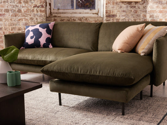 Olive green sectional sofa with large cushions against an exposed brick wall. Decor includes a pink patterned pillow and neutral-toned pillows.