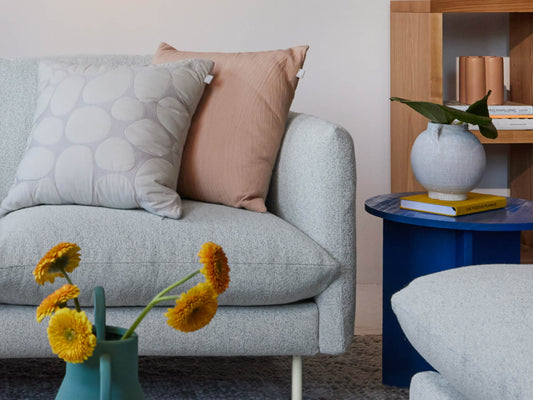 Light grey sofa with two cushions, one grey and one beige. Nearby, a blue side table with a vase and books. Yellow flowers in a teal vase.