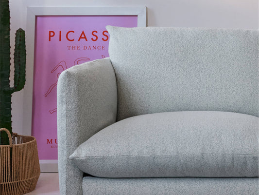 Light grey sofa with textured upholstery, next to a framed pink artwork featuring "Picasso." A cactus in a wicker basket sits beside it.