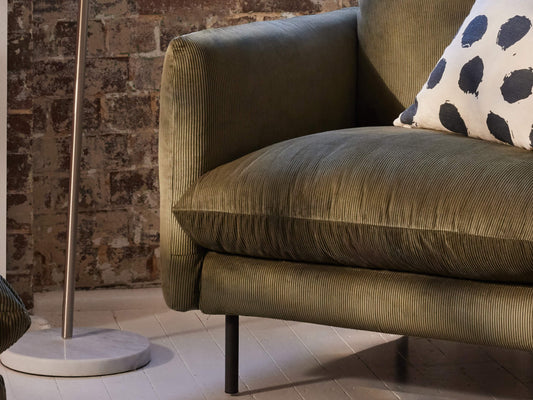 Green corduroy sofa with a white cushion featuring black spots, set against an exposed brick wall. A sleek floor lamp is nearby.