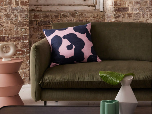 Green sofa with a pink and navy patterned cushion, next to a pink side table. Brick wall backdrop and decorative vases on the table.